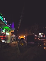 A dimly lit urban street scene at night featuring a bank with illuminated green signage on the left. There are several people walking along the sidewalk, motorcycles parked, and a vehicle on the road. Streetlights and vehicle headlights provide minimal lighting, casting shadows and creating a moody atmosphere.