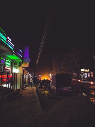A dimly lit urban street scene at night featuring a bank with illuminated green signage on the left. There are several people walking along the sidewalk, motorcycles parked, and a vehicle on the road. Streetlights and vehicle headlights provide minimal lighting, casting shadows and creating a moody atmosphere.