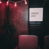 A dimly lit room features a sign reading 'Laidback Lover' on a dark wall. The ambiance is enhanced by string lights with red bulbs. A potted plant is seen in the corner, and plush chairs with a muted color are positioned below the sign.