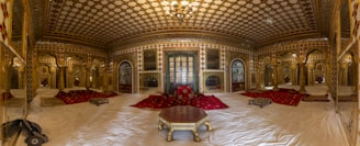 A lavish and opulent room with intricate patterns and golden accents. The ceiling and walls are adorned with detailed geometric designs, mostly in red, gold, and white. The room features luxurious red carpets and cushioned seating areas, with a central octagonal table. The overall decor suggests a royal or historical ambiance.