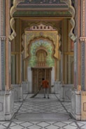 A person is sweeping the floor in a beautifully decorated corridor with intricate designs and vibrant colors. The corridor is detailed with floral and geometric patterns, and there is an ornate door at the end, surrounded by traditional Indian architecture elements.