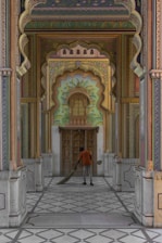 A person is sweeping the floor in a beautifully decorated corridor with intricate designs and vibrant colors. The corridor is detailed with floral and geometric patterns, and there is an ornate door at the end, surrounded by traditional Indian architecture elements.