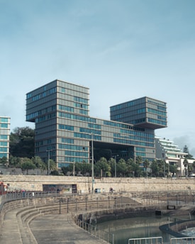 A modern architectural structure featuring interconnected rectangular buildings with numerous windows. The buildings are surrounded by greenery and stand near a stone embankment with steps leading down to a waterway in the foreground.