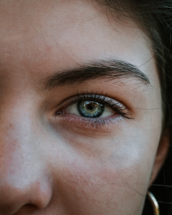 A close-up of a human eye with green and blue hues, surrounded by eyelashes and a well-defined eyebrow. The skin around the eye shows natural texture and some freckles. The lighting highlights the eye's roundness and creates a reflection within the iris.