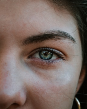A close-up of a human eye with green and blue hues, surrounded by eyelashes and a well-defined eyebrow. The skin around the eye shows natural texture and some freckles. The lighting highlights the eye's roundness and creates a reflection within the iris.