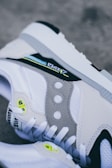 A close-up view of a sneaker highlighting its design elements such as the gray suede panel with circular cutouts, neon green eyelets, and detailed stitching. The white and black accents are prominent, creating a modern and stylish look.
