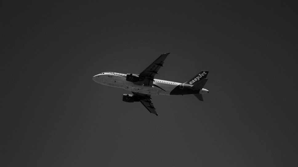 A black and white photograph of an airplane flying against a dark sky. The aircraft is seen from a low angle with the wings and tail visible. The word 'easyJet' is prominently displayed on the tail.