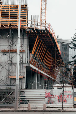 A construction site featuring a partially-built structure with exposed scaffolding and metal beams. The area is enclosed with a temporary barrier featuring images of construction workers and the word 'TEAMSWORK'. A tall crane is visible in the background. Sparse trees line the pavement in front of the construction area.