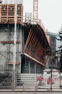 A construction site featuring a partially-built structure with exposed scaffolding and metal beams. The area is enclosed with a temporary barrier featuring images of construction workers and the word 'TEAMSWORK'. A tall crane is visible in the background. Sparse trees line the pavement in front of the construction area.