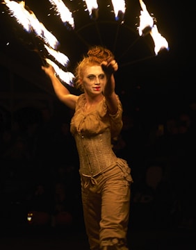 A performer is skillfully manipulating a fan with multiple fire torches against a dark background. The individual is dressed in a vintage-style beige outfit, with elaborate makeup and hair styled in an updo. The scene captures dynamic movement and the warmth of the firelight accentuates the performer, highlighting a dramatic and captivating performance.