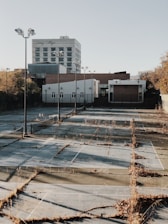 A deserted tennis court with overgrown weeds between the cracks of the surface. Several tall light poles are positioned along the court, but the nets are missing. In the background, there is a multi-story building labeled 'Laney College' and some smaller buildings surrounded by a fence. The scene is bathed in warm, late afternoon sunlight.