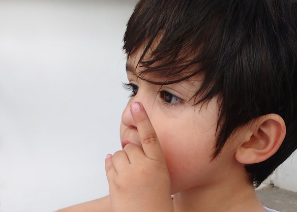 A young child with dark hair is viewed from the side. The child is touching their nose with their index finger, appearing thoughtful or curious. The background is a soft, off-white color, which highlights the child's facial features.