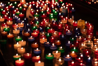 A large number of small candles are arranged closely together, each encased in a colorful holder. The candle holders are in various colors, including red, blue, green, and clear, creating a vivid and warm scene as the candle flames flicker gently.