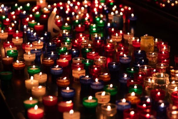 A large number of small candles are arranged closely together, each encased in a colorful holder. The candle holders are in various colors, including red, blue, green, and clear, creating a vivid and warm scene as the candle flames flicker gently.
