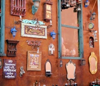 A rustic wall adorned with various vintage and eclectic decorations, including mirrors, lanterns, signs, a clock, and artwork. The wall is painted in a warm, earthy tone with accents of turquoise and blue. There is a humorous sign stating, 'NO tenemos WI-FI, HABLEN entre ustedes.'