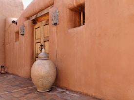 A large, smooth, tan clay pot with intricate designs sits in front of a matching adobe-style wall. The wall features a wooden door with metal accents and decorative elements. The paved ground is made of brick, with muted earth tones creating a warm atmosphere.