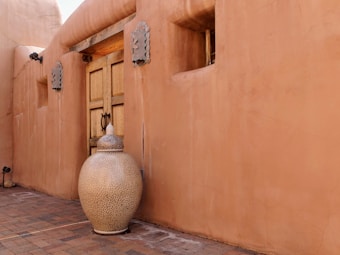 A large, smooth, tan clay pot with intricate designs sits in front of a matching adobe-style wall. The wall features a wooden door with metal accents and decorative elements. The paved ground is made of brick, with muted earth tones creating a warm atmosphere.