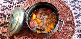 A metal cooking pot filled with a rich stew containing large pieces of beef and vegetables like tomatoes and peppers. The pot, accompanied by its lid, rests on a woven placemat with a colorful geometric pattern.