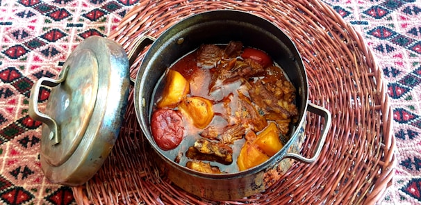 A metal cooking pot filled with a rich stew containing large pieces of beef and vegetables like tomatoes and peppers. The pot, accompanied by its lid, rests on a woven placemat with a colorful geometric pattern.