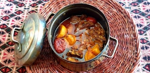 A metal cooking pot filled with a rich stew containing large pieces of beef and vegetables like tomatoes and peppers. The pot, accompanied by its lid, rests on a woven placemat with a colorful geometric pattern.