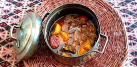 A metal cooking pot filled with a rich stew containing large pieces of beef and vegetables like tomatoes and peppers. The pot, accompanied by its lid, rests on a woven placemat with a colorful geometric pattern.