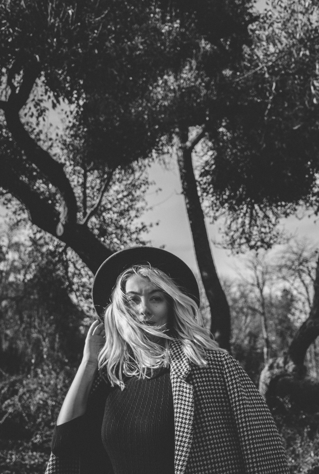 grayscale photo of woman with hat standing near tree