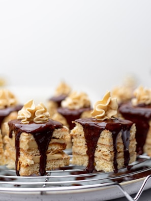Small layered cakes are topped with smooth chocolate ganache and a swirl of cream. The cakes consist of stacked layers, showcasing their moist crumb and creamy filling. They are arranged on a cooling rack against a soft, blurred background.