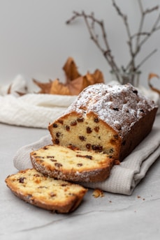 A loaf of fruitcake is displayed on a light-colored cloth. The cake is sliced, revealing a golden interior with scattered raisins. Powdered sugar lightly dusts the top of the loaf. In the background, blurred autumn leaves and twigs add a seasonal touch to the setting.