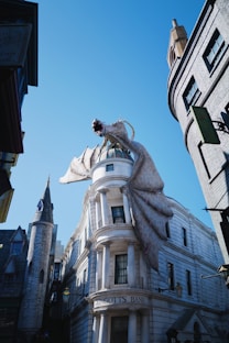 A fantasy setting features a large dragon perched atop Gringotts Bank with its mouth open, positioned among tall buildings under a clear blue sky. The dragon's wings are spread, adding a dramatic flair to the street scene.