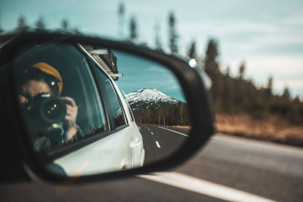 close-up photography of woman taking photo of mountain through vehicle side mirror