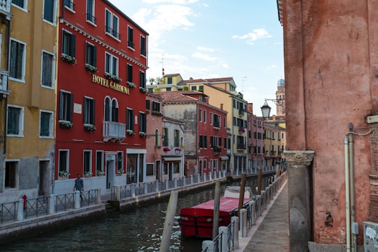 A picturesque canal scene featuring a series of colorful buildings with arched windows and decorative flower boxes. The red building has the words 'Hotel Gardena' displayed on it. The canal is bordered by a narrow walkway and lined with poles, and a small boat with a red cover is floating in the water. The sky is partly cloudy, adding a serene atmosphere.