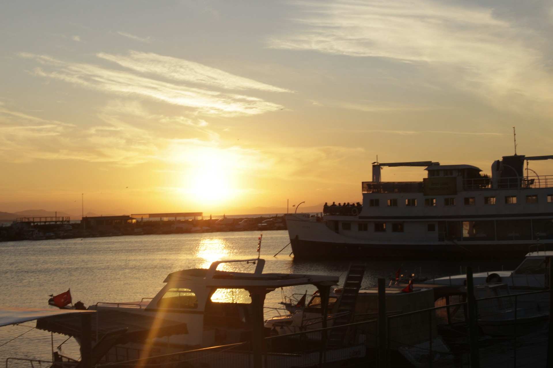 A scenic view of the Port Jefferson ferry dock at sunset, with soft light reflecting on the water and a hint of the pizzeria’s light red and yellow signage in the foreground.