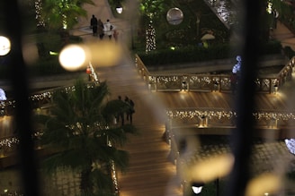 A nighttime scene depicting a park or garden area with illuminated paths and railings adorned with string lights. A few people are walking along the wooden walkway, surrounded by lush greenery and palm trees. Spherical light fixtures add a soft, warm glow to the environment, enhancing the festive atmosphere.