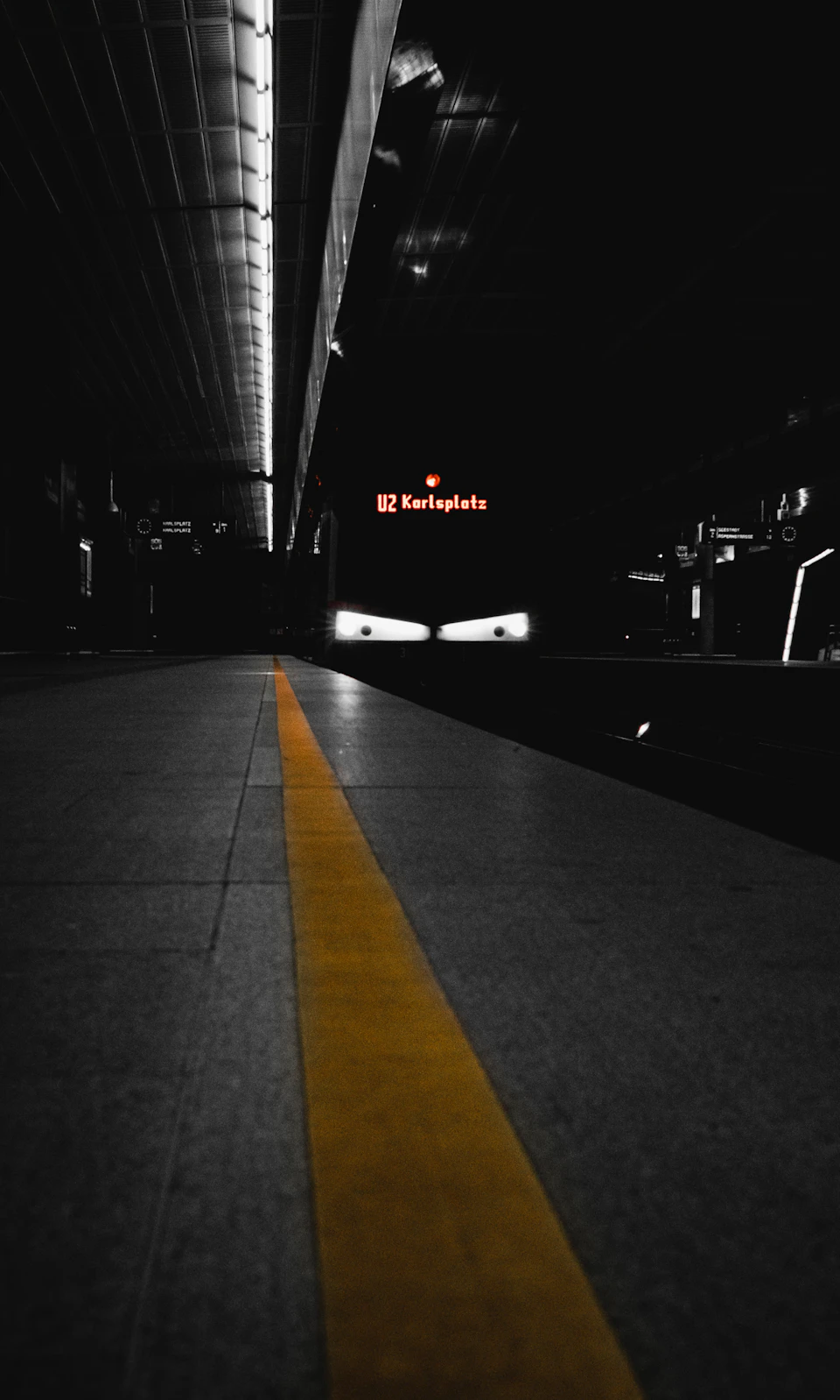 The image depicts a train station at night, with a yellow line running down the center of the platform and a large sign reading "Kilmholt" on one side. The platform is illuminated by overhead lights, creating a stark contrast against the darkness outside. A few blurred figures can be seen in the background, adding depth to the scene.