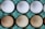 A dozen eggs arranged in a 3x4 pattern sit in a blue egg carton. The eggs vary in color from white to tan, each occupying a separate compartment in the textured carton.