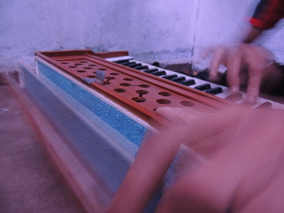 A motion-blurred image featuring hands playing a harmonium, with focus on the keyboard and air holes. The harmonium is primarily wooden with some blue accents.