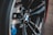 Close-up view of a car wheel featuring a BMW logo prominently displayed at the center, surrounded by sleek, shiny black spokes and hints of blue components in the background.