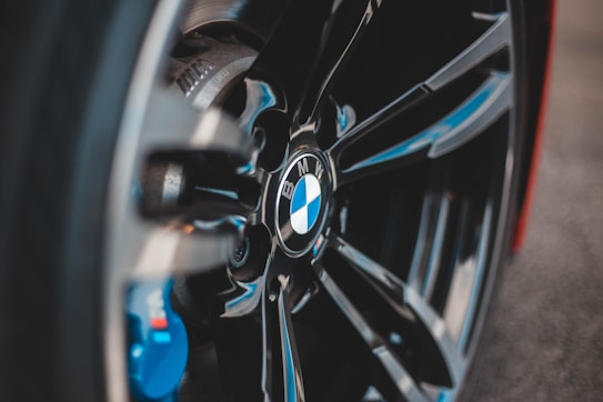 Close-up view of a car wheel featuring a BMW logo prominently displayed at the center, surrounded by sleek, shiny black spokes and hints of blue components in the background.