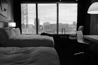 A hotel room features two neatly made beds with white linens. A large window with blinds provides a view of a cityscape, including buildings and a crane. The room is furnished with a desk, a chair, and a standing lamp. A suitcase is positioned next to the window.