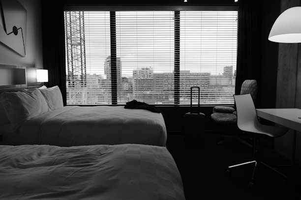 A hotel room features two neatly made beds with white linens. A large window with blinds provides a view of a cityscape, including buildings and a crane. The room is furnished with a desk, a chair, and a standing lamp. A suitcase is positioned next to the window.