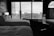 A hotel room features two neatly made beds with white linens. A large window with blinds provides a view of a cityscape, including buildings and a crane. The room is furnished with a desk, a chair, and a standing lamp. A suitcase is positioned next to the window.