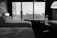 A hotel room features two neatly made beds with white linens. A large window with blinds provides a view of a cityscape, including buildings and a crane. The room is furnished with a desk, a chair, and a standing lamp. A suitcase is positioned next to the window.