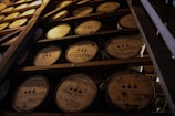 Rows of oak barrels aging bourbon in a dimly lit warehouse with rustic wooden beams.