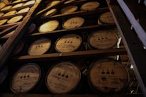 Several wooden barrels stacked in a dimly lit storage area, each barrel marked with labels indicating they contain bourbon whiskey. The atmosphere is rustic and industrial, with visible wooden beams supporting the structure.