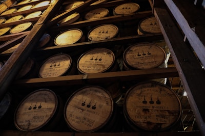 Several wooden barrels stacked in a dimly lit storage area, each barrel marked with labels indicating they contain bourbon whiskey. The atmosphere is rustic and industrial, with visible wooden beams supporting the structure.