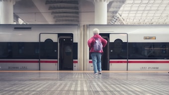 A person with short white hair stands on a train platform facing a modern train stopped at the station. The person is wearing a red jacket and a purple backpack, with blue jeans. The train doors are open, and the station features a high ceiling with geometric designs.