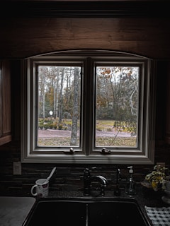 A kitchen sink with a black countertop is positioned below a double window. Outside the window, there is a view of a garden with trees and foliage displaying autumn colors. Inside, a coffee mug with a spoon, a soap dispenser, and a small plant are on the countertop.