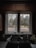 A kitchen sink with a black countertop is positioned below a double window. Outside the window, there is a view of a garden with trees and foliage displaying autumn colors. Inside, a coffee mug with a spoon, a soap dispenser, and a small plant are on the countertop.
