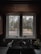 A kitchen sink with a black countertop is positioned below a double window. Outside the window, there is a view of a garden with trees and foliage displaying autumn colors. Inside, a coffee mug with a spoon, a soap dispenser, and a small plant are on the countertop.