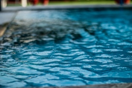 A close-up view of a pool with clear blue water, rippling gently across the surface. The background appears slightly blurred with hints of greenery and possibly poolside furniture.
